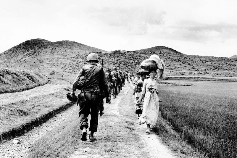 A line of US Army soldiers file past a group of Korean women and children during the Korean War